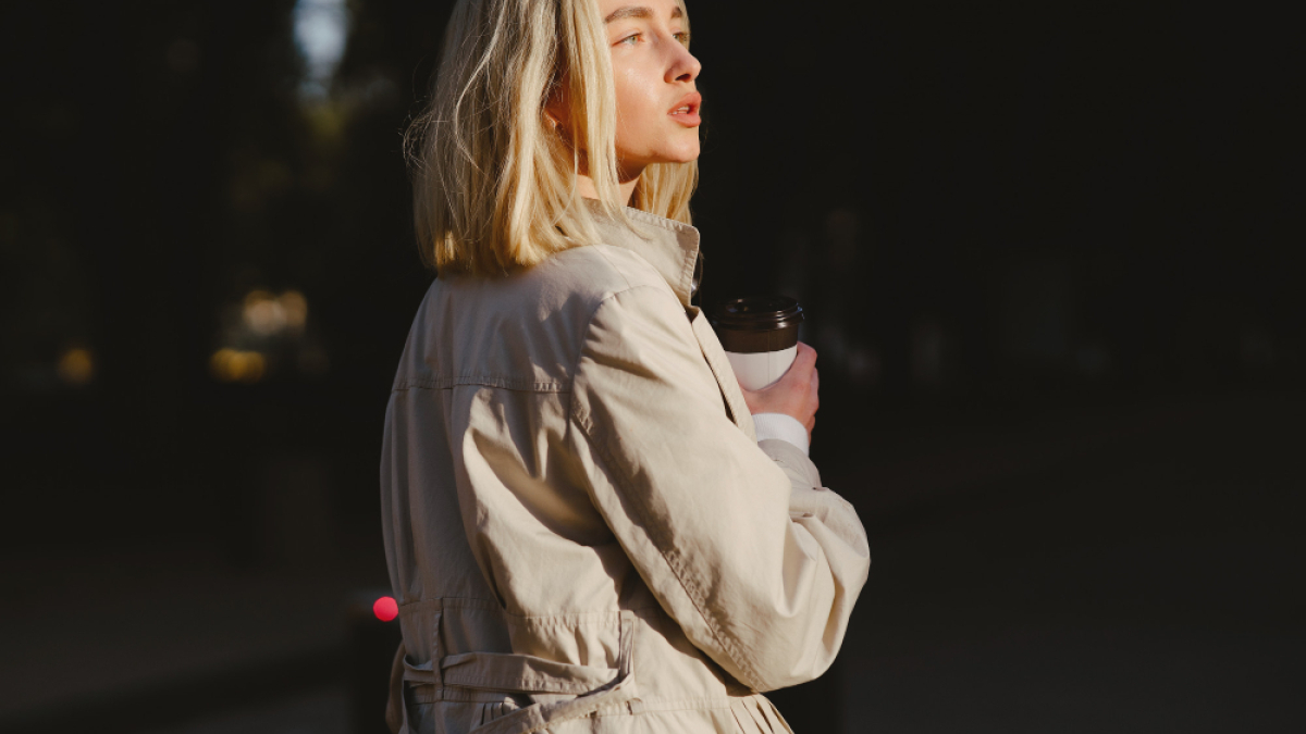 Девушка в плаще со стаканчиком кофе на размытом фоне.