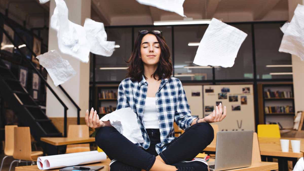 Девушка в клечатой рубашке занимается йогой, сидя на столе в офисе, а вокруг нее летает бумага