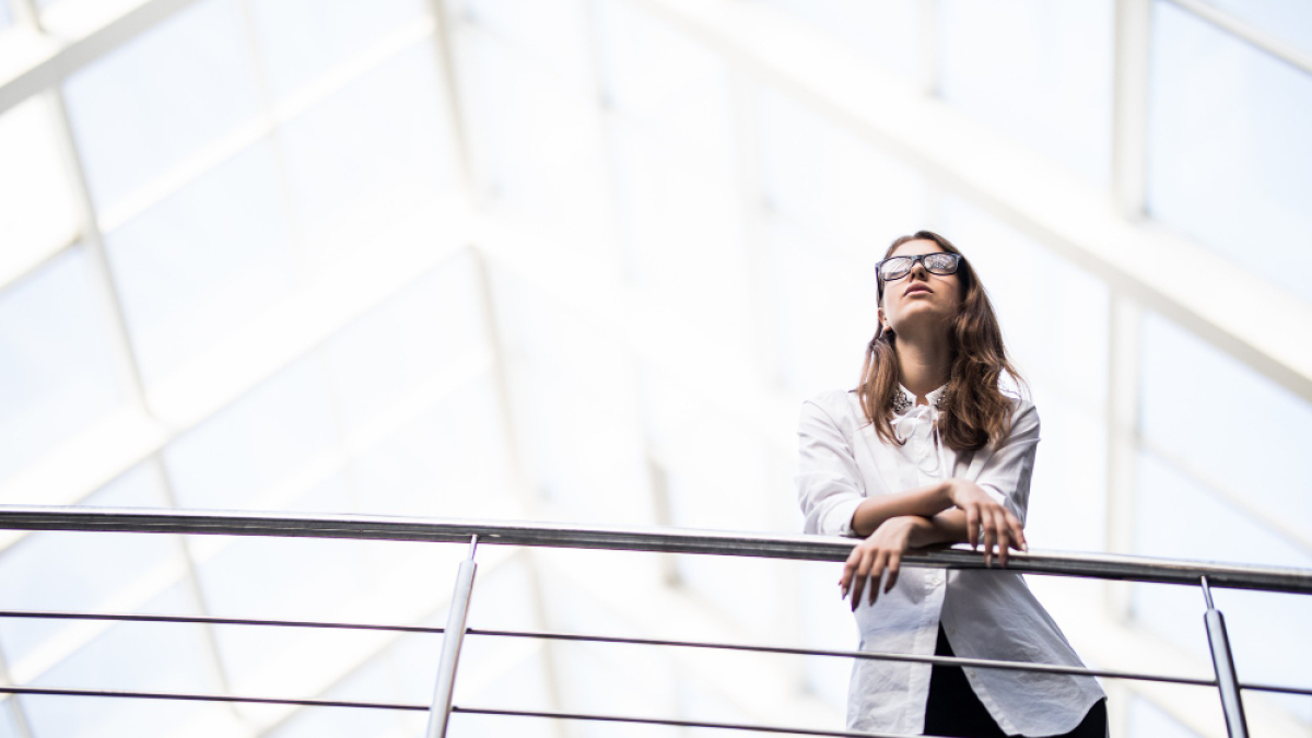 Девушка в очках стоит под стеклянной крышей, опираясь на перила.