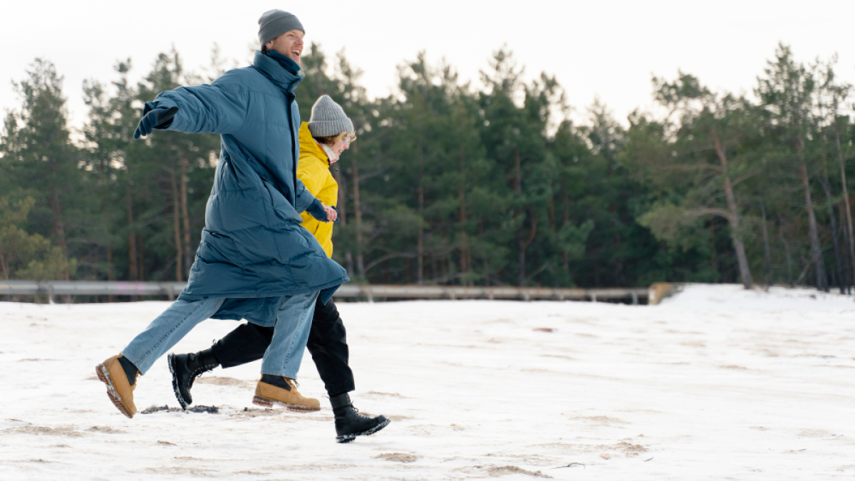 Мужчина и женщина в теплой одежде держатся за руку и бегут по снегу.