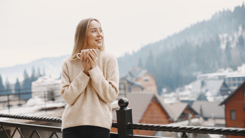 Девушка в свитере стоит на балконе на фоне гор и держит чашку в руках.
