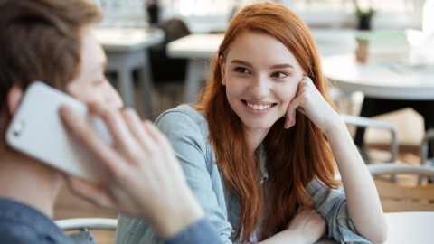 Рыжая девушка с улыбкой смотрит на молодого человека, который говорит по телефону.