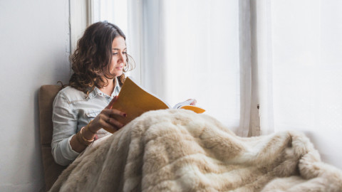 Девушка читает книгу у окна, укутавшись в плед