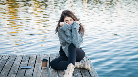 Девушка в свитере сидит у воды, рядом лежит телефон и стоит стаканчик с кофе.