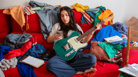 Деввушка с гитарой и телефоном сидит на заваленном вещами диване.